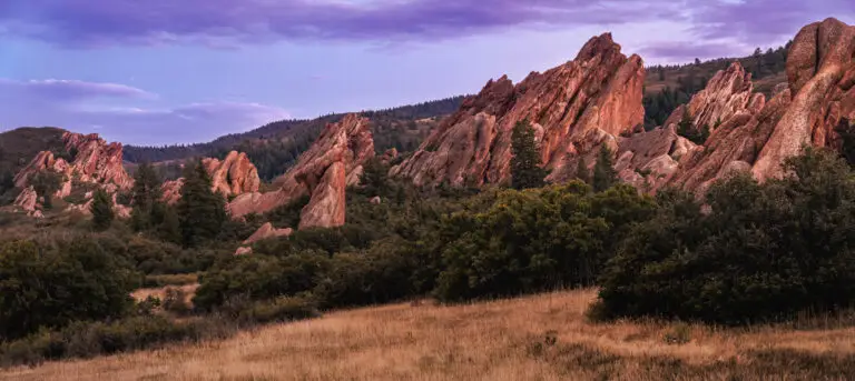 Jagged red rock formations rise over a forested plain at dusk with a purple sky.