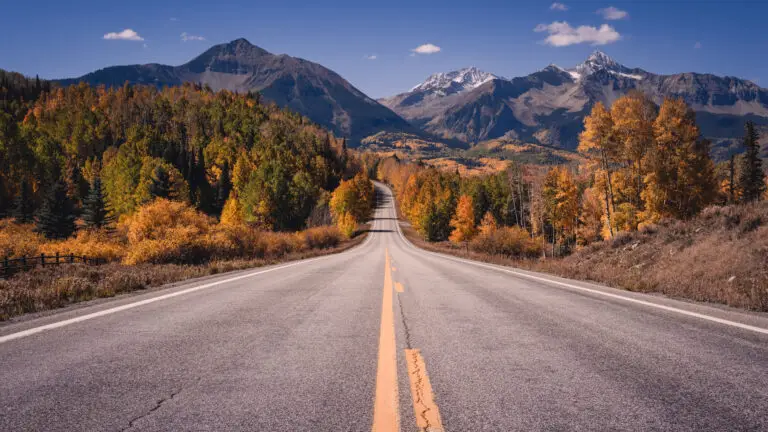 Two-lane road through autumn forest toward snow-capped mountains under a blue sky