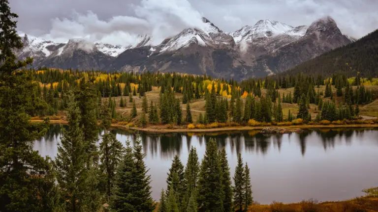 Colorado Photography Locations Colorado Photography Locations Snow capped mountains rise behind a forest of evergreen trees with gold autumn foliage reflected in a calm lake at the shore