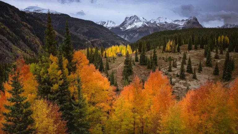 Colorado Photography Locations Colorado Photography Locations Autumn forest in a valley with orange yellow and green trees a small wooden cabin and snowcapped mountains in the distance