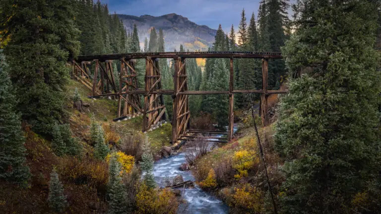 Wooden railroad trestle bridge spans a rushing river, framed by a dense evergreen forest and distant mountains.