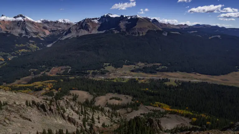 Aerial view of a mountain valley with snow‑capped peaks, dense evergreen forest, and open meadows under a blue sky.
