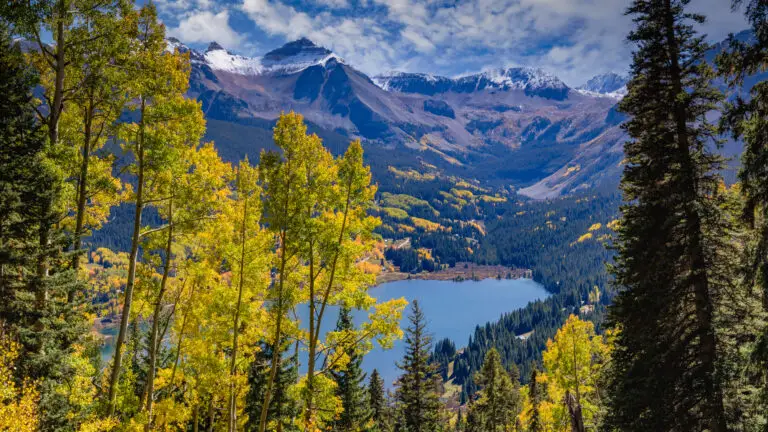 Snowy mountain range over a forested valley with a blue lake, framed by golden aspen trees in autumn.