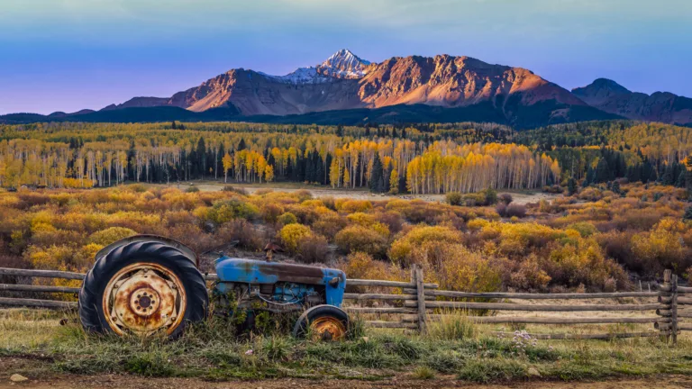 Rusty blue tractor beside a wooden fence in a fall landscape with colorful trees and distant mountains, some snow on peaks.