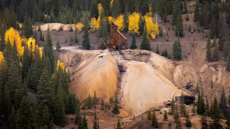Rusty wooden mining shack perched on a sandy hill amid autumn trees with yellow leaves.